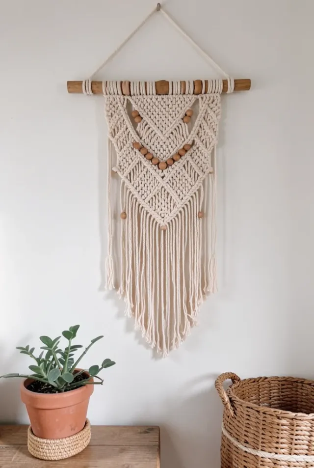 Intricate macrame wall hanging with fringe and wooden beads above a potted plant and woven basket