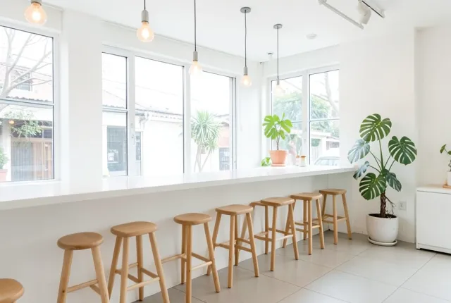 Bright minimalist cafe interior with long white counter, wooden stools, large window, and potted monstera