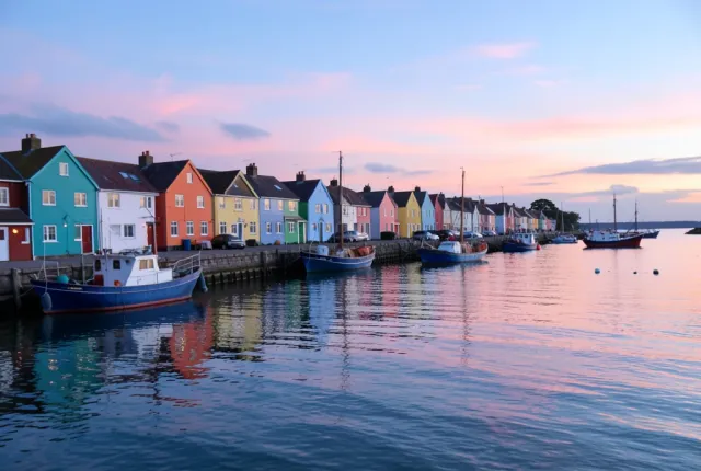 Colorful painted cottages along a quiet harbor with fishing boats and a pink-blue sunrise sky