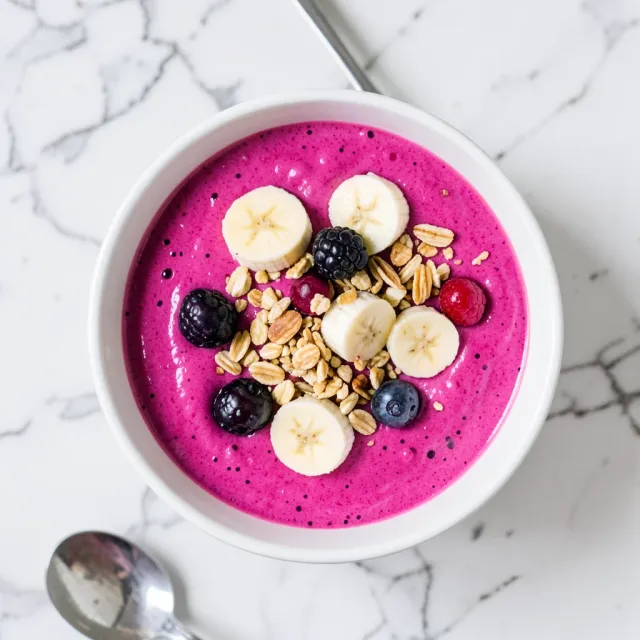 Overhead view of pink acai smoothie bowl topped with banana, granola, and berries on marble