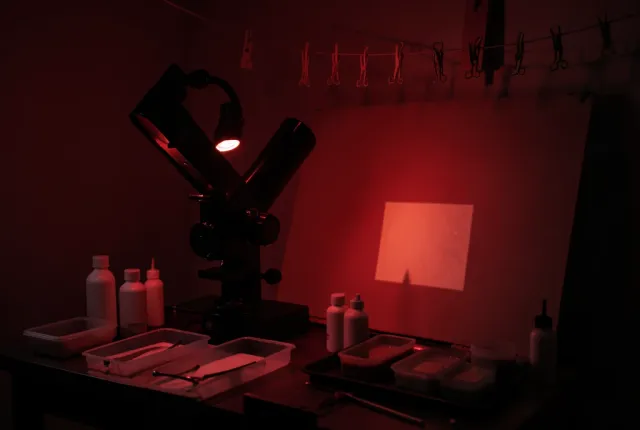 Red-safelight darkroom with enlarger projecting onto an easel, chemical trays and blank bottles