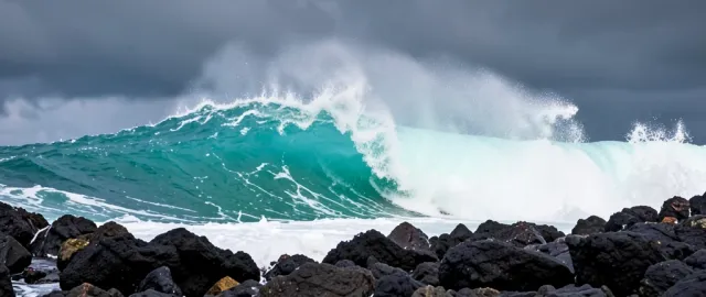 Huge turquoise ocean wave breaking against dark volcanic rocks with white spray and stormy sky