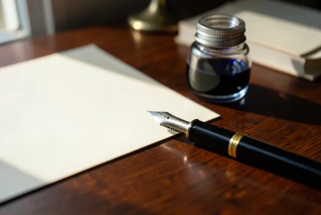 Uncapped fountain pen with gold nib beside a blue-black inkwell on cream paper, soft window light