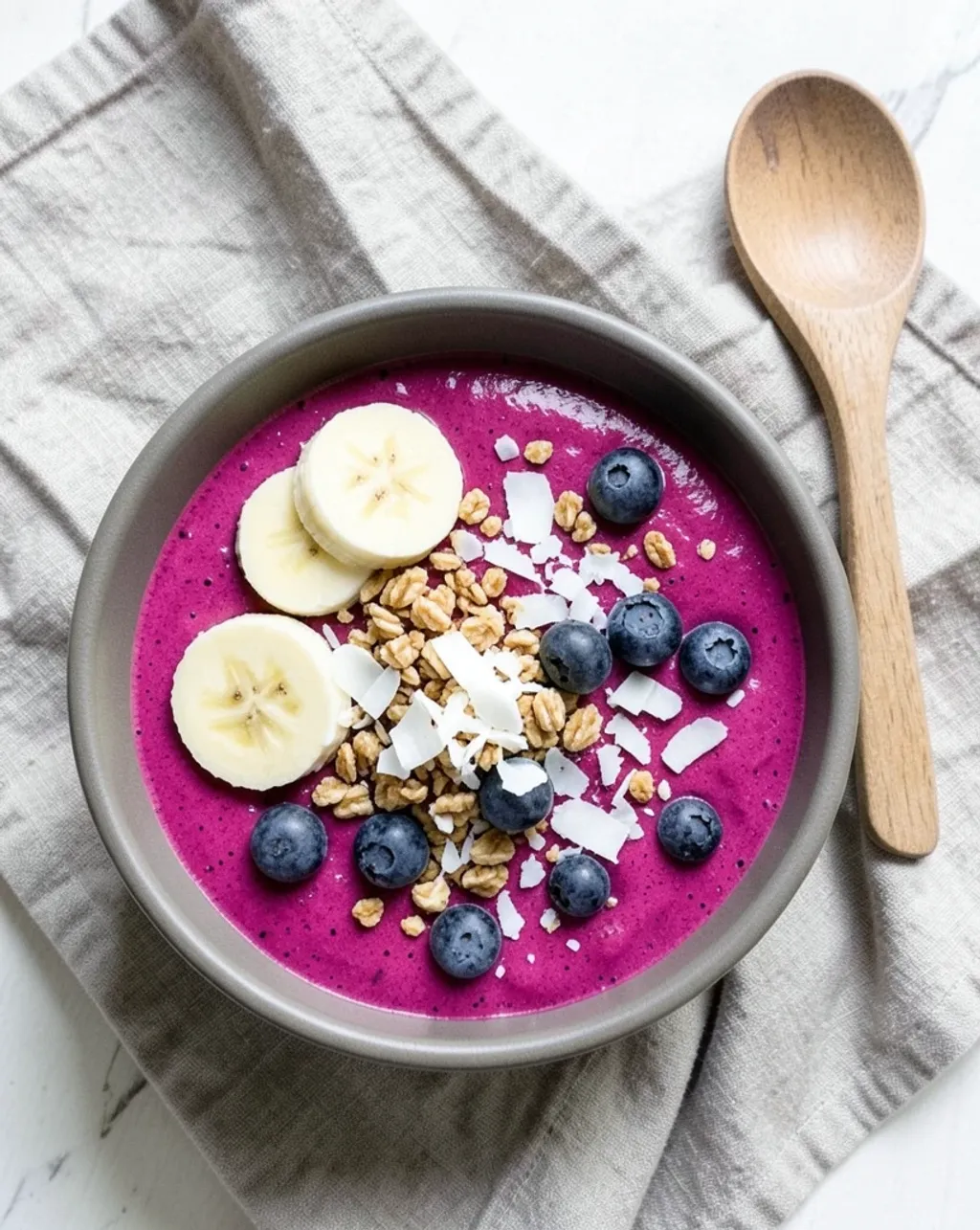 Overhead flat lay of a magenta acai smoothie bowl with banana, blueberries, granola, and coconut flakes