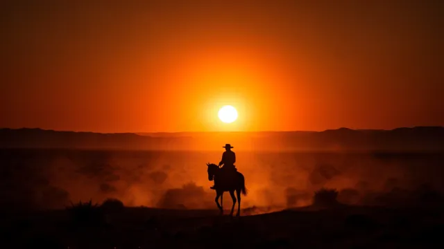 Silhouette of a lone cowboy on horseback against a dramatic orange sunset over a wide desert landscape
