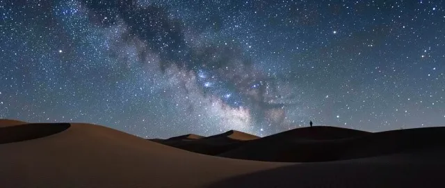 Sweeping sand dunes under a bright Milky Way arc with a lone figure on a distant ridge