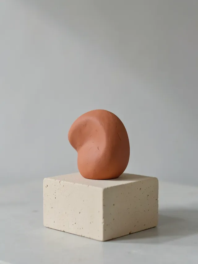 Minimal abstract terracotta clay sculpture on a beige pedestal in soft studio light