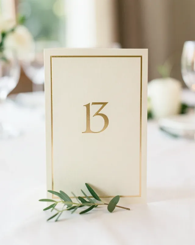 Elegant ivory wedding table number card with a thin gold frame and eucalyptus sprig on a soft-lit reception table