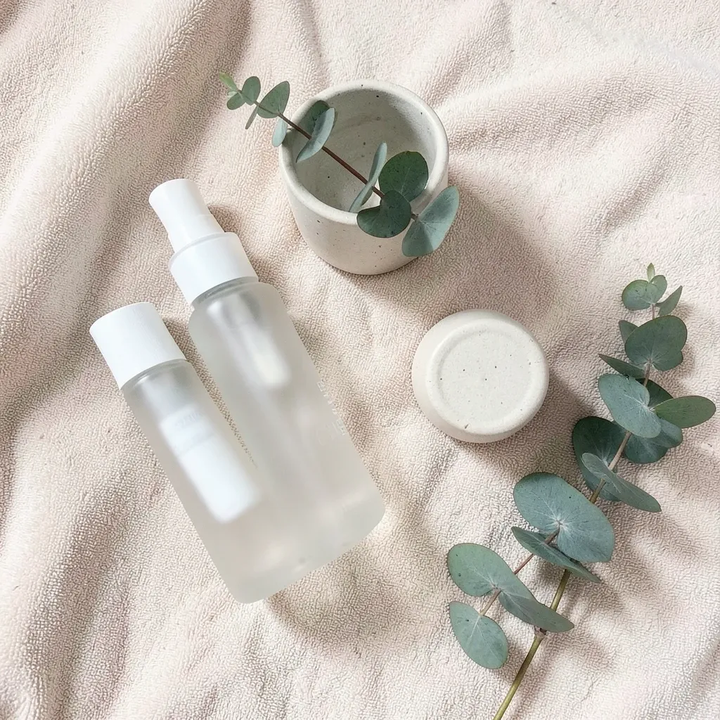 Minimalist skincare flat lay with frosted bottles, ceramic jar, eucalyptus on beige towel