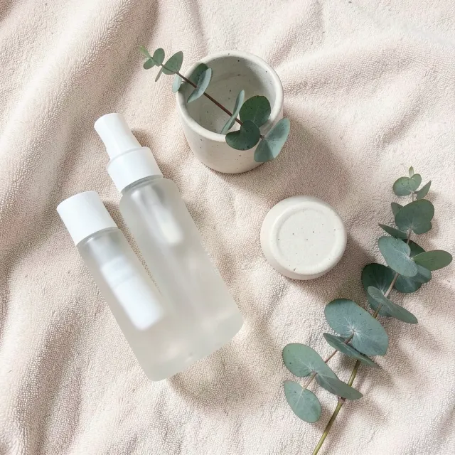 Minimalist skincare flat lay with frosted bottles, ceramic jar, eucalyptus on beige towel