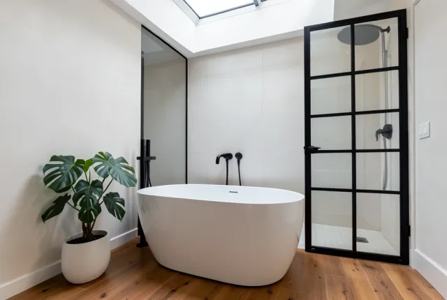 Modern bathroom with white freestanding tub, black shower screen, monstera plant, and skylight