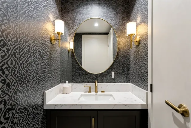 Modern powder room with black patterned wallpaper, brass sconces, round mirror, and marble countertop