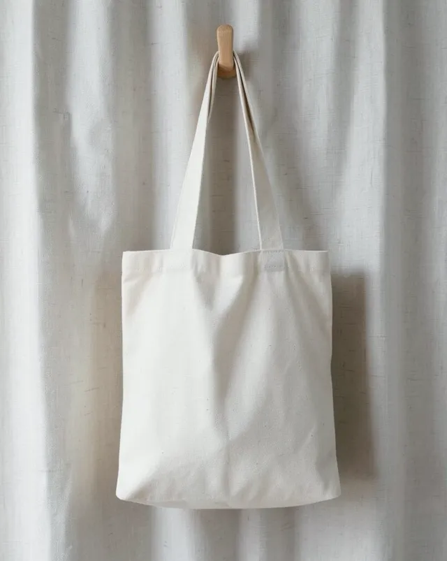 Plain canvas tote bag with blank front panel hanging on a wood hook against linen curtain