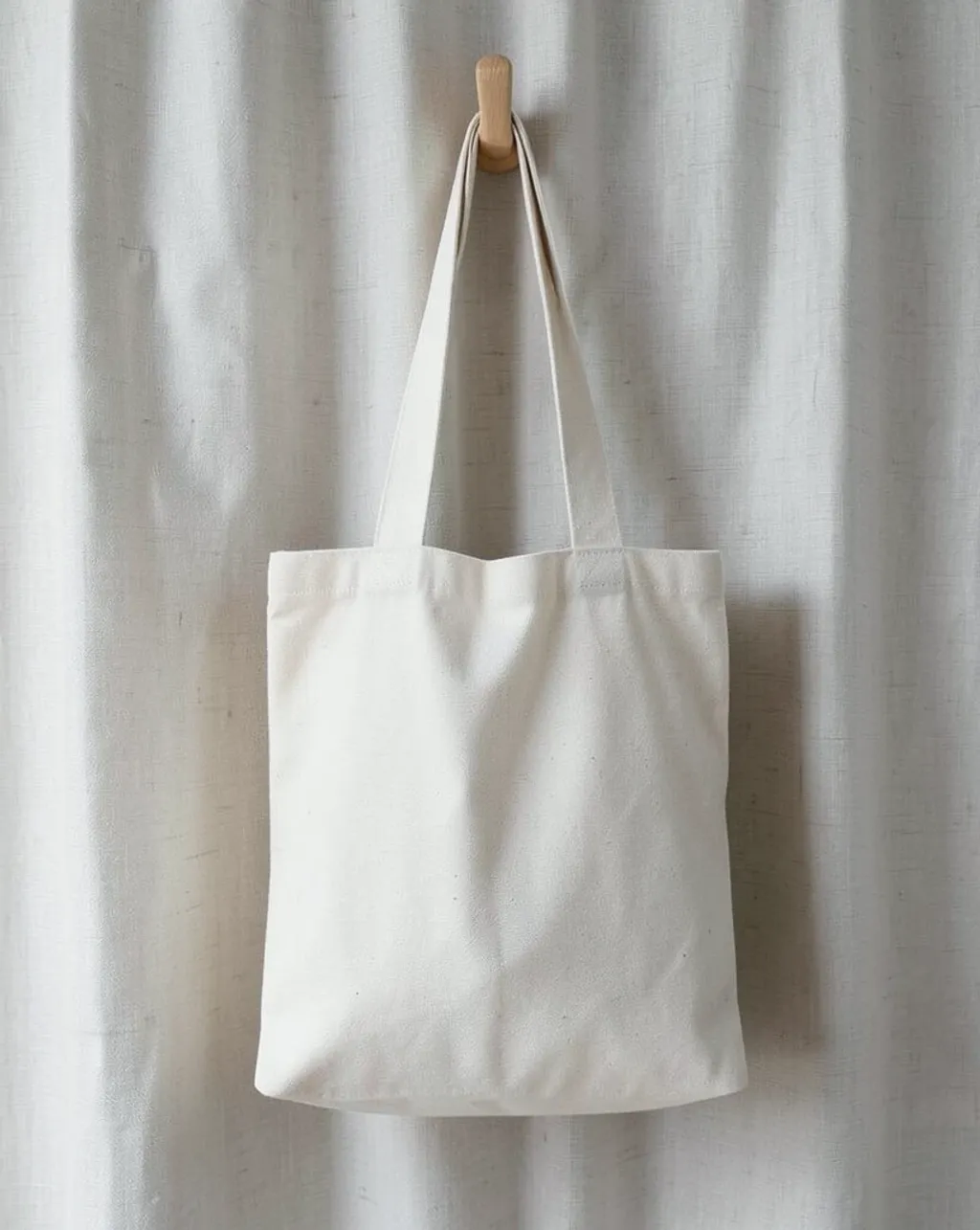 Plain canvas tote bag with blank front panel hanging on a wood hook against linen curtain