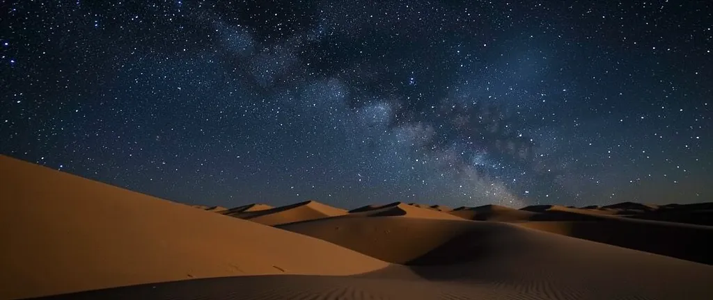 Sweeping Sahara sand dunes under a vast starry night sky with soft moonlight shadows