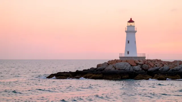 White lighthouse on a rocky point with calm sea and soft pink and orange sunrise sky