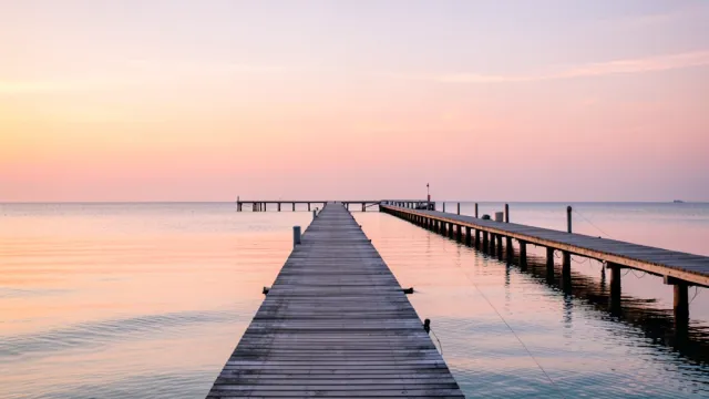 Long wooden pier stretching into calm ocean with pink and orange sunrise reflected in glassy water