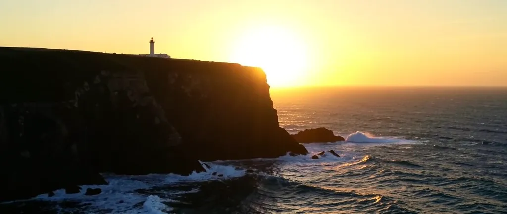 Golden sunrise behind dark coastal cliffs with crashing waves and distant lighthouse