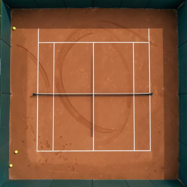 Overhead view of a terracotta clay tennis court with white lines, net shadow and broom arcs