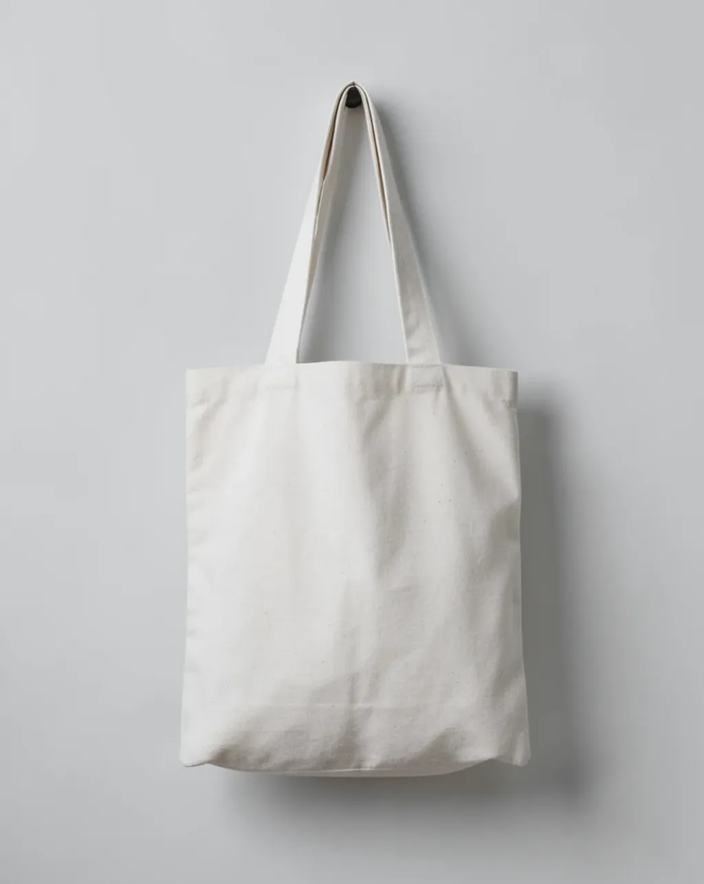 A blank natural canvas tote bag hanging on a grey wall with empty front space
