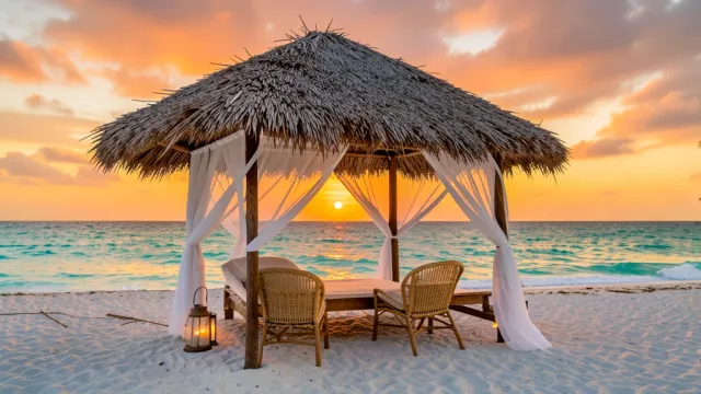 Thatched beach cabana with blowing sheer curtains, woven chairs, turquoise ocean, and orange sunset sky