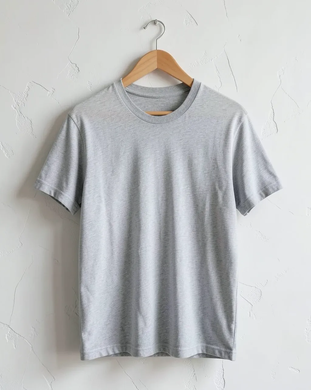 Blank heather-grey t-shirt on a wooden hanger against a plaster wall in soft natural light