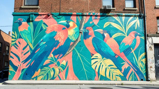 Vibrant tropical bird and jungle leaf mural painted on a sunlit brick city wall
