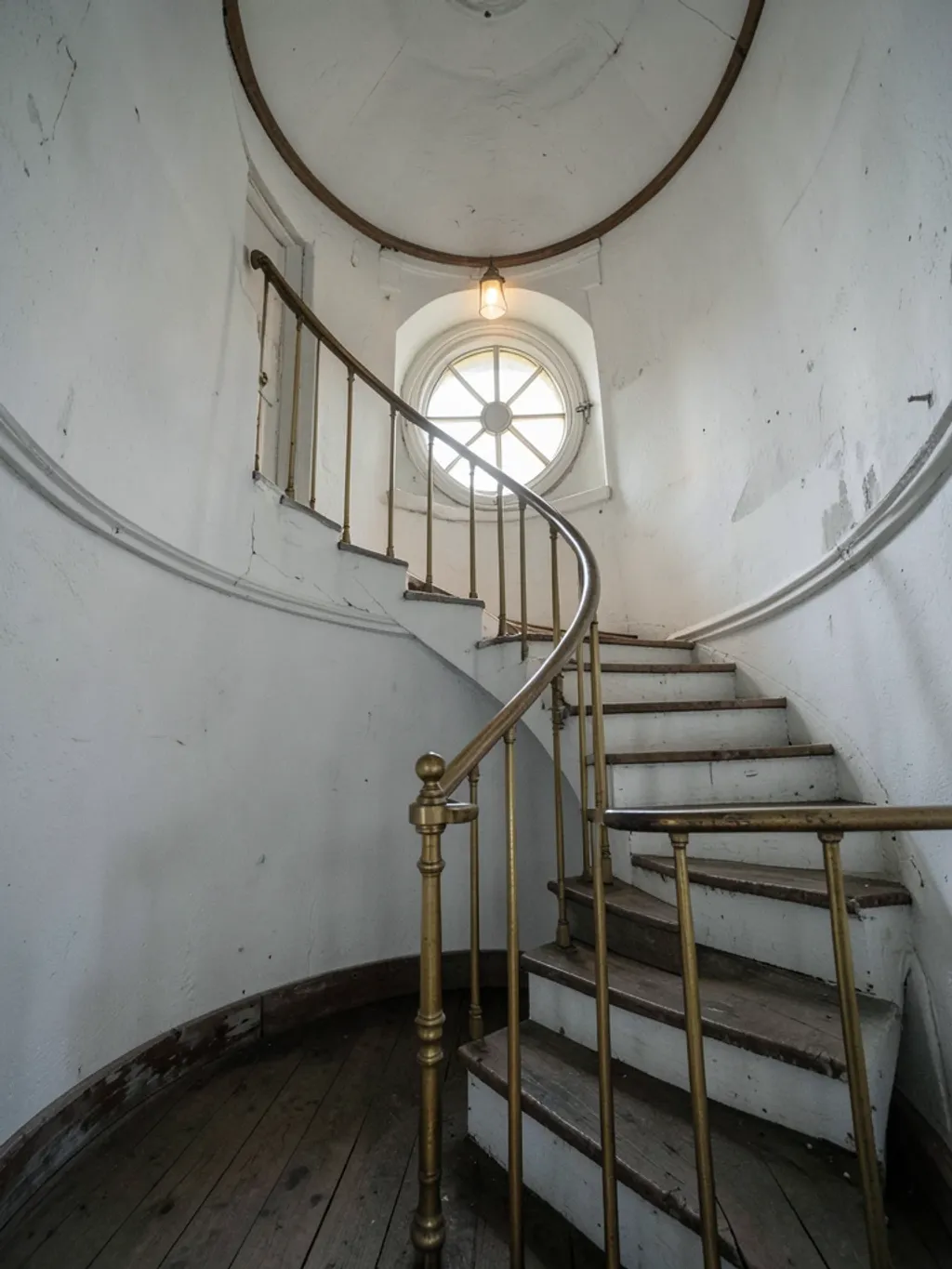 Round white-washed lighthouse interior with spiral staircase, brass railing, and glowing Fresnel lens