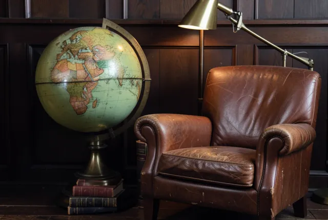 Antique world globe beside a worn leather armchair with books, brass lamp, and dark wood paneling