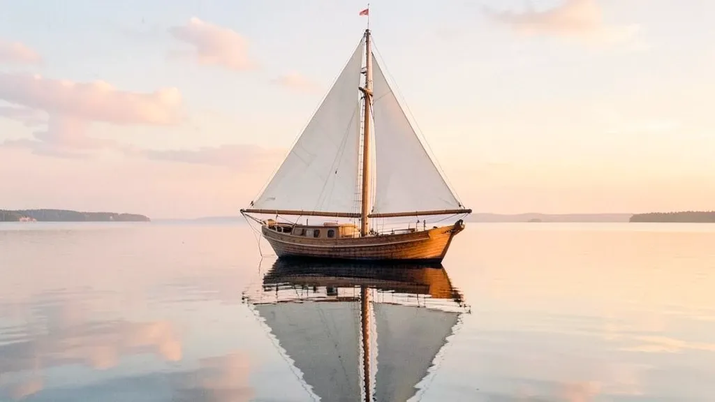Classic wooden sailboat with white sails reflected in calm water under a pastel sunrise sky